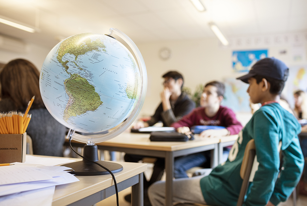 En bild ur ett klassrum där en jordglob är i förgrunden och elever i bakgrunden sittandes vid sina platser. 